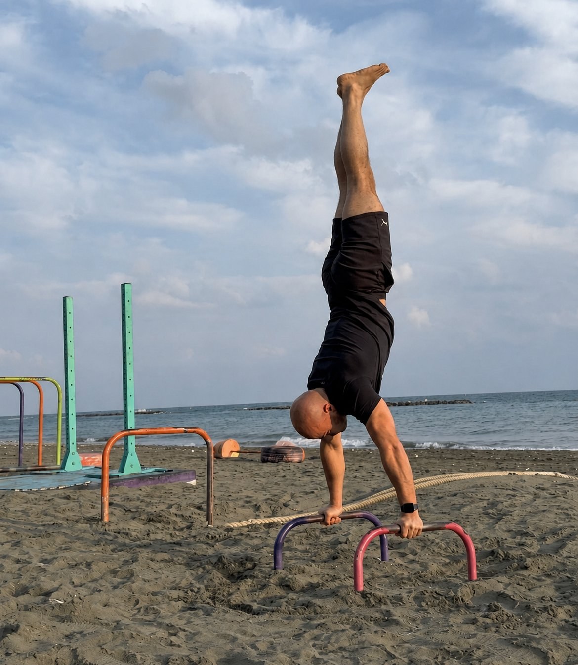 Ruben performing a handstand on parallettes at an outdoor beach training area in Limassol.